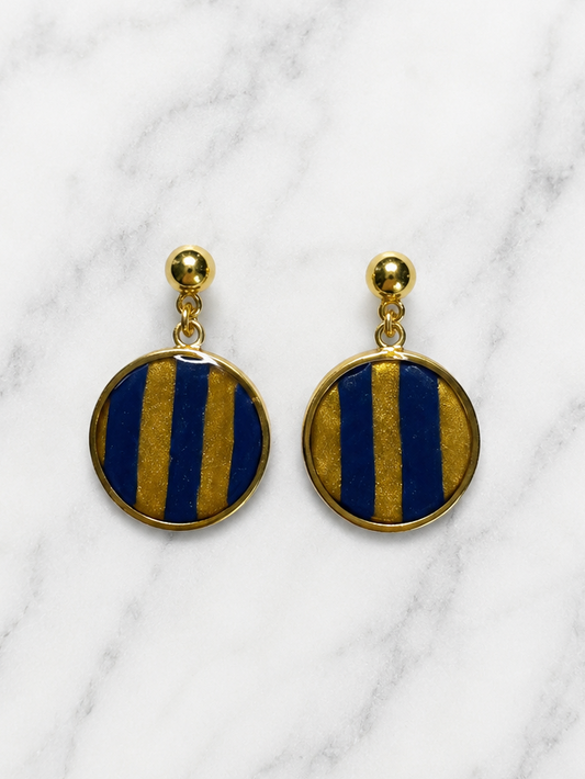 A pair of gold-tone drop earrings with circular blue and gold striped enamel faces, displayed on a white marble background.